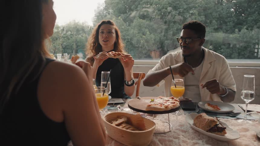 Diverse friends enjoy a cheerful lunch, eating pizza and chatting happily at a beautifully lit restaurant table