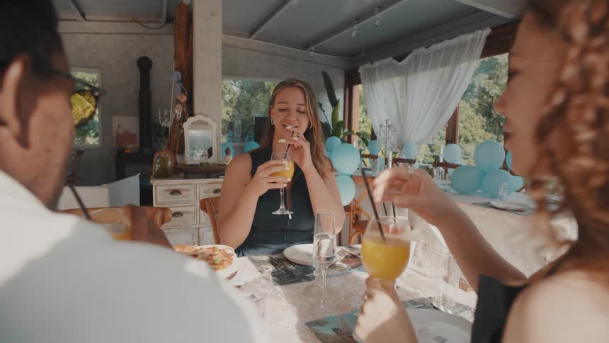 Happy friends make a toast with their drinks while sitting at a decorated table in a sunlit restaurant