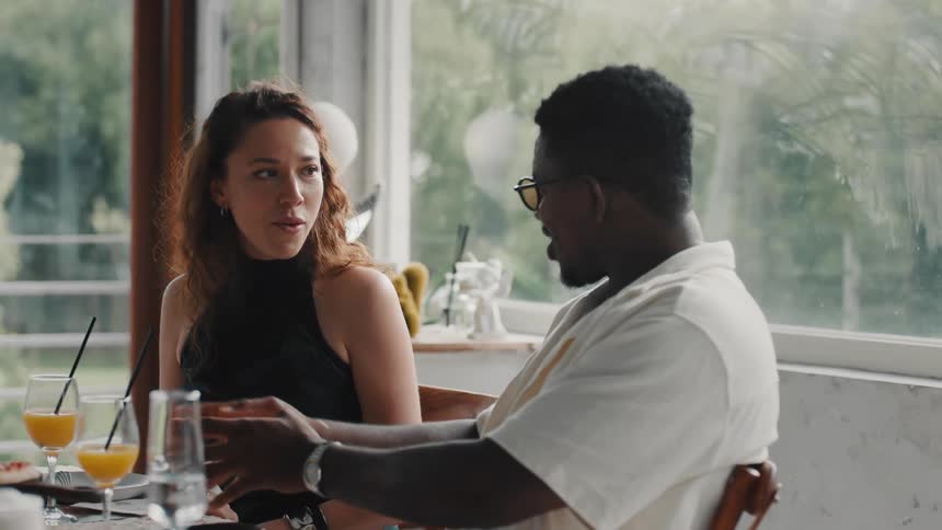 An expressive young woman and man share a lively and animated conversation over drinks and food in a beautiful, light-filled restaurant