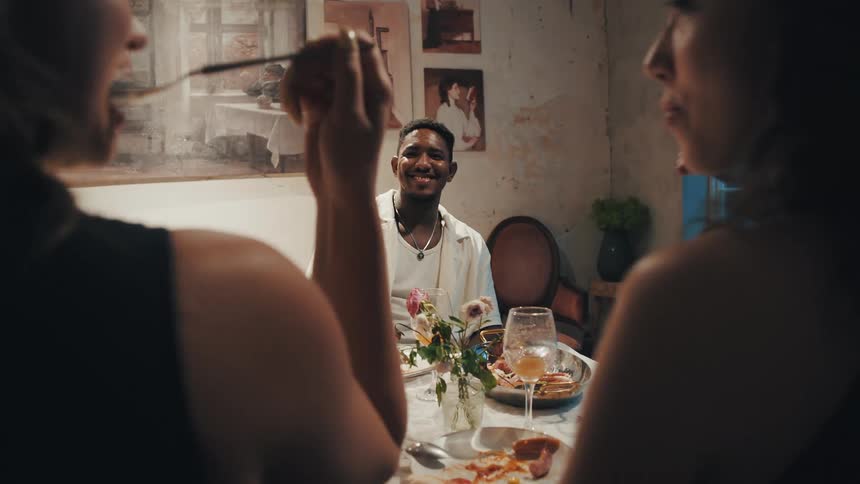 A happy young man smiles at his friends over dinner in a warmly lit, artistic room