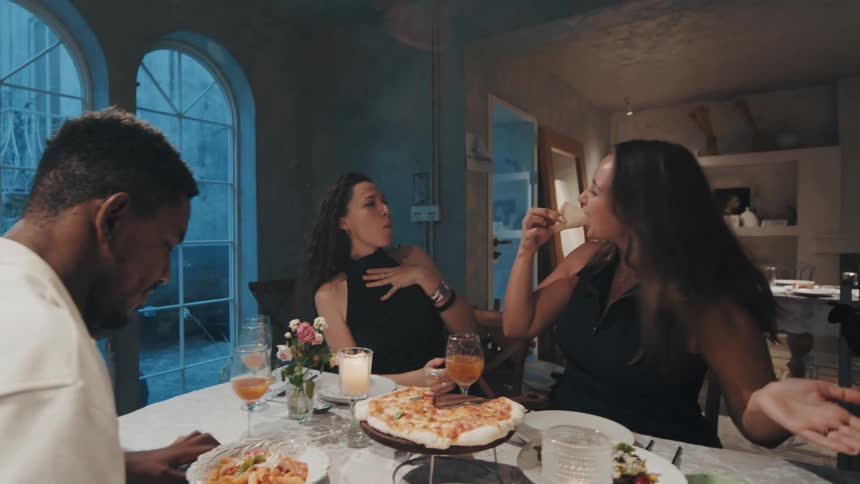 In slow motion, a woman playfully feeds her friend a slice of pizza during a lively and fun dinner party with another friend