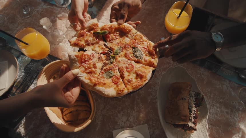 Top-down slow-motion view of friends' hands taking cheesy slices of pizza from a sunlit restaurant table