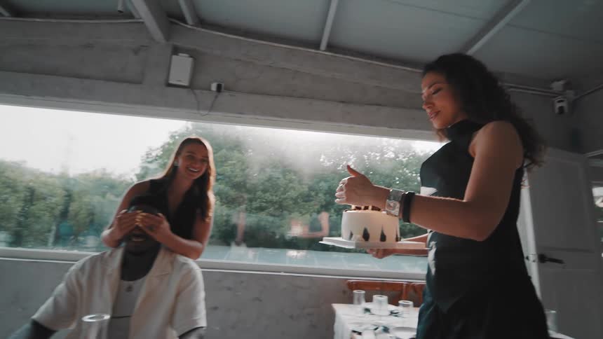 A young woman brings a beautiful birthday cake with lit candles to surprise her friend at a sunlit restaurant