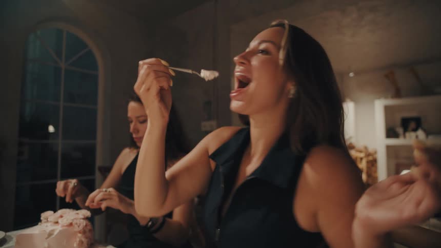 A happy woman playfully eats a spoonful of pink frosted cake while celebrating with a friend at an evening party