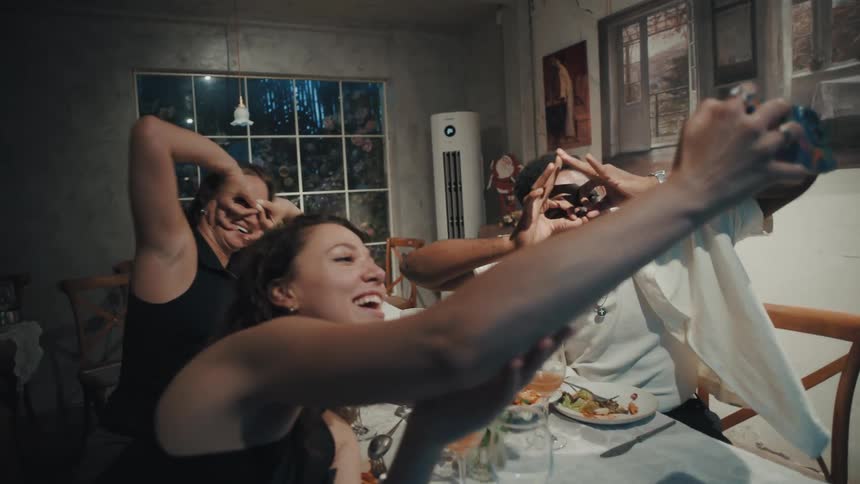 A playful group of multicultural friends laugh and make silly faces while taking a selfie together at a dinner party