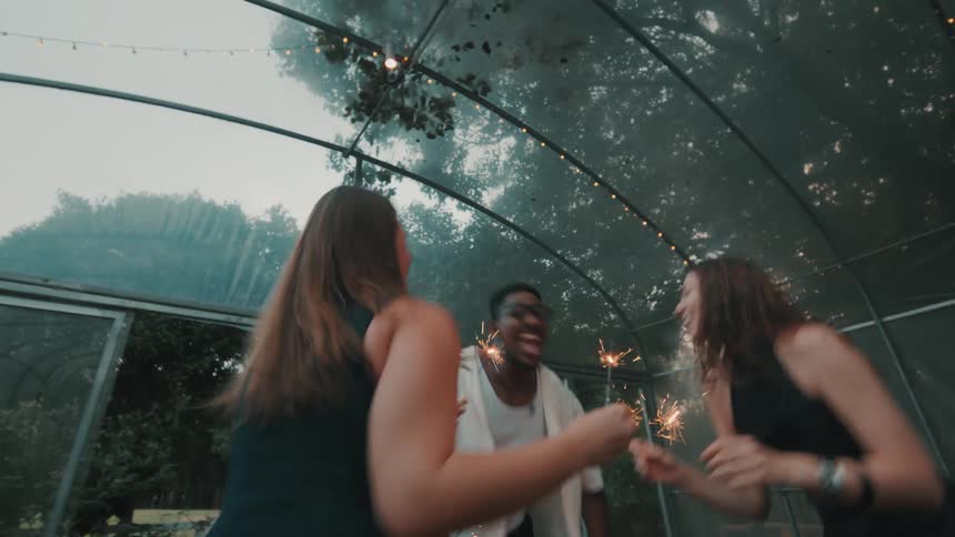 Joyful multicultural friends dance energetically with burning sparklers at a party inside a greenhouse