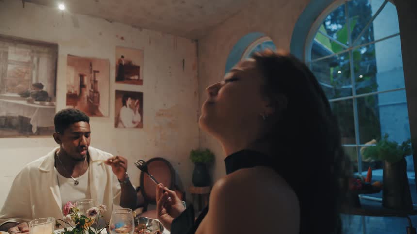 A woman savors a bite of food from a fork in slow motion while dining with friends in an artistic room