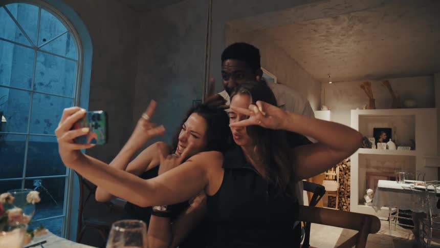 A diverse group of friends laugh and make funny faces while taking a selfie together at a stylish dinner party