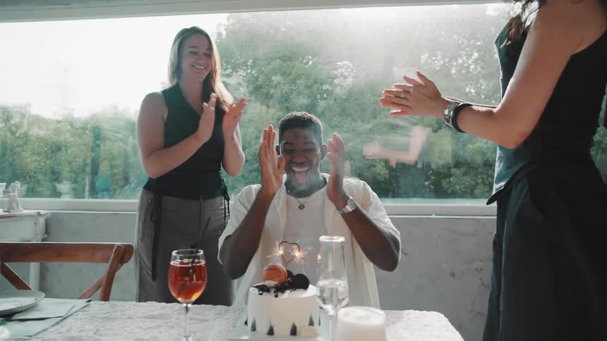 Two joyful young women surprise their male friend with a birthday cake and sparkler in a sunlit restaurant, leading to a happy celebration and hugs