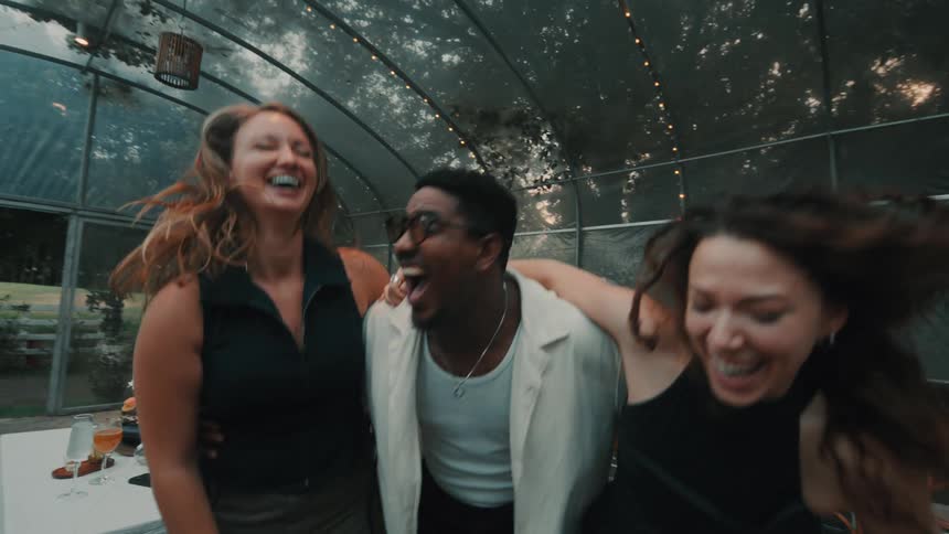 A diverse group of friends laugh and dance energetically in slow motion inside a beautiful greenhouse party tent at dusk