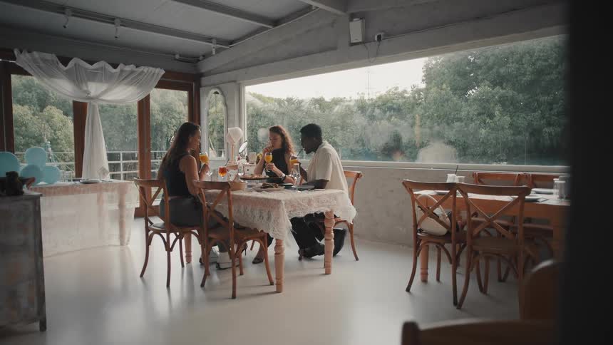 Three diverse friends enjoy a pleasant conversation and toast with orange juice while having brunch in a stylish restaurant with large windows
