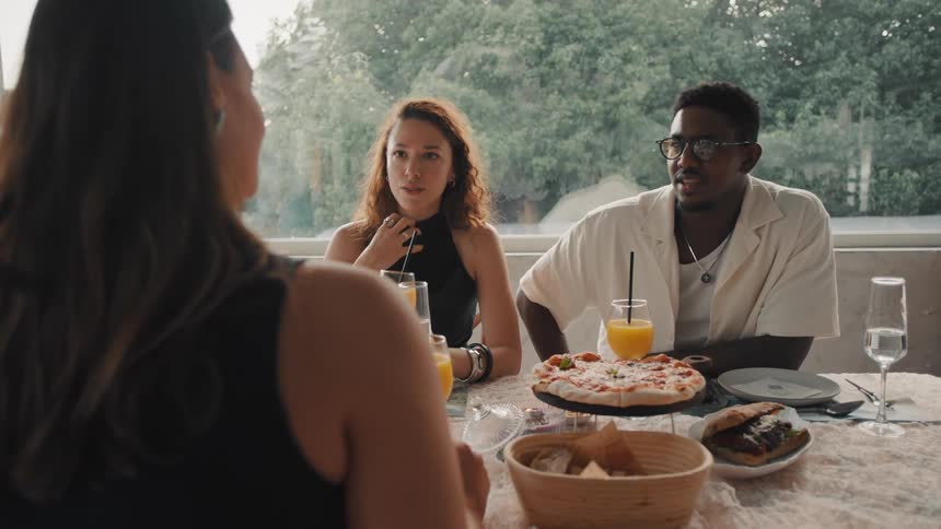 Diverse group of friends laugh and chat while enjoying pizza and juice at a restaurant table with a bright window view