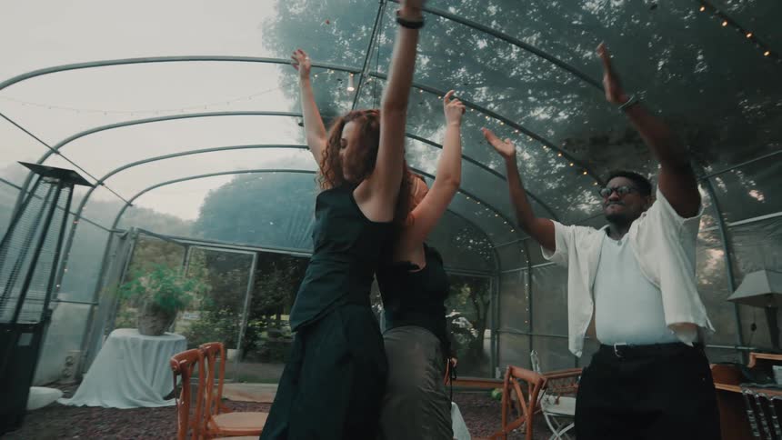 A diverse group of friends dance joyfully in slow motion at a party inside a greenhouse decorated with string lights