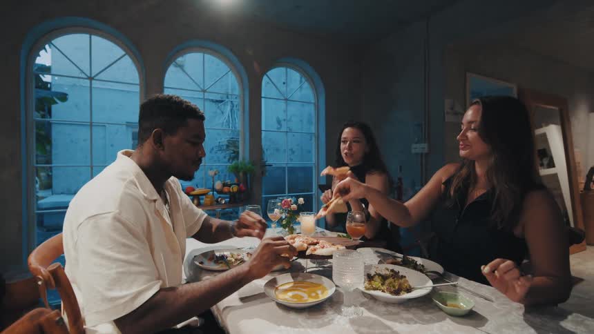 A diverse group of friends happily eats pizza and has fun together during a dinner party at home