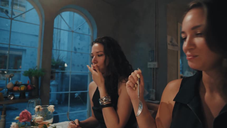 In slow motion, a woman playfully feeds her laughing friend a bite of food during a fancy evening dinner