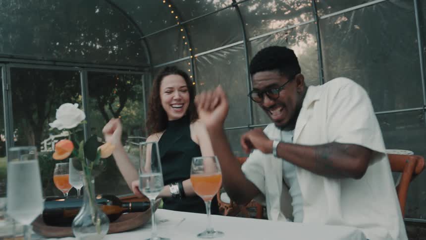 A group of happy, diverse friends laugh and dance together while seated at a dinner party inside a beautiful greenhouse