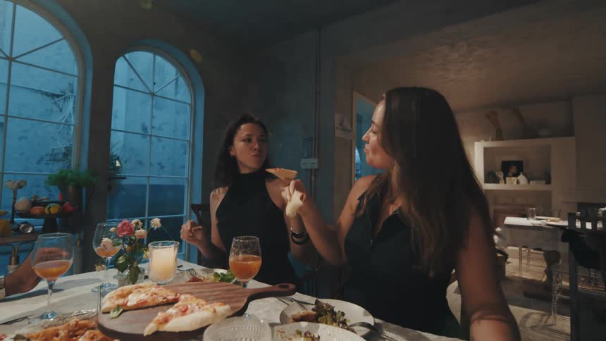 Joyful friends laugh and playfully toast with pizza slices in slow motion during a fun dinner party at a stylish restaurant