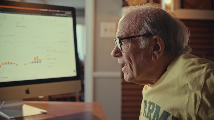 An elderly man reacts with excitement and surprise while analyzing data charts on his computer at his home desk