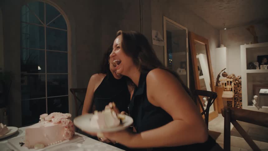 Two cheerful women burst into laughter while one playfully feeds the other a slice of birthday cake during a dinner celebration