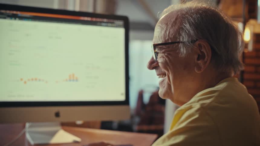 Happy senior man with glasses smiles while analyzing financial charts and data on his desktop computer