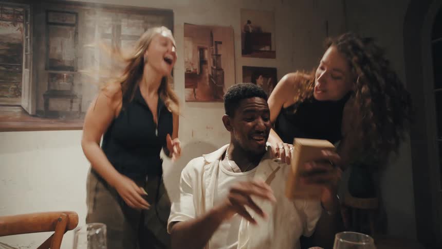 A man reacts with joyful surprise while opening a birthday gift as his two female friends cheer and dance excitedly around him in slow motion