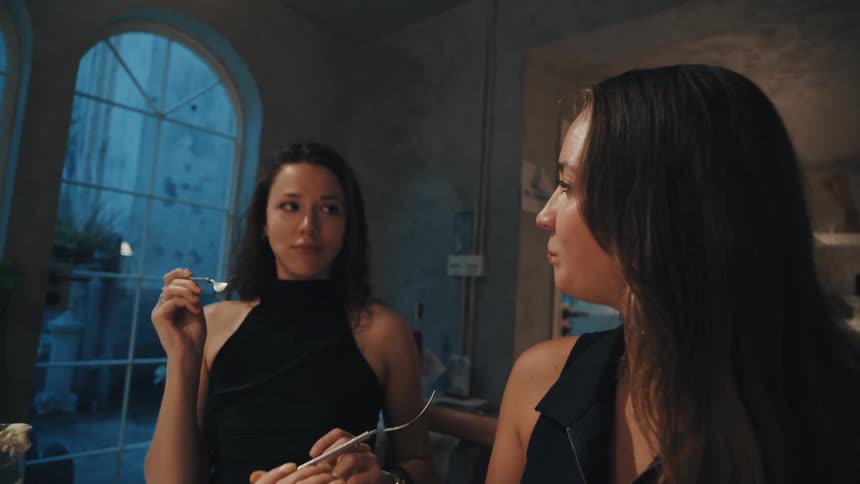 A woman playfully feeds her friend a bite of food from her fork as they enjoy dinner together in a stylish restaurant