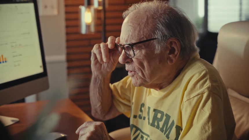An elderly U.S. Army veteran takes off his glasses and reacts with shock while looking at financial data on his computer at home