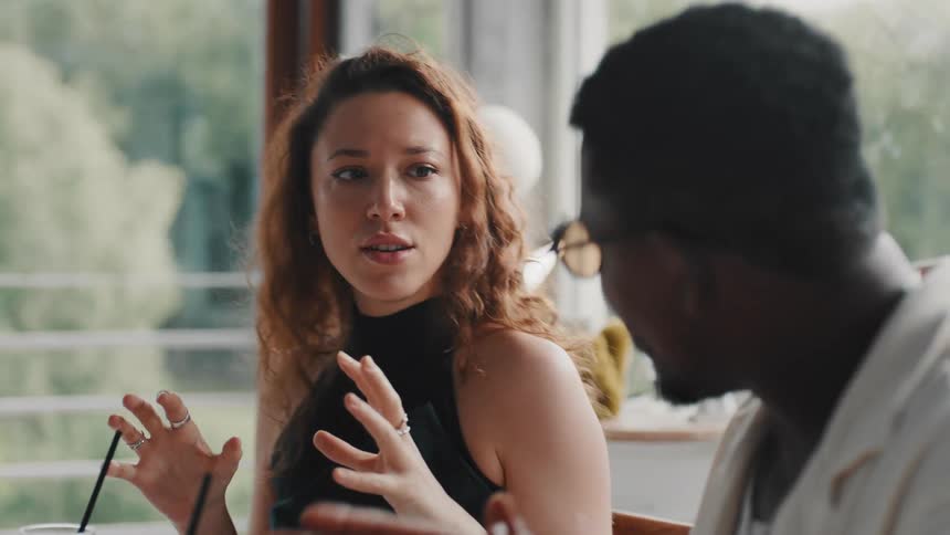 A beautiful young woman with curly hair animatedly converses with a man at a restaurant table during the day