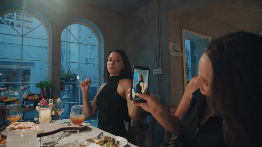 A woman records her friend dramatically eating pasta and dancing at the dinner table for a social media video