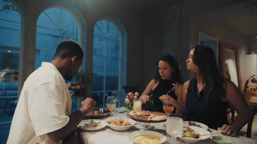 A diverse group of friends enjoys a meal and conversation together at a cozy, candlelit dinner party at night