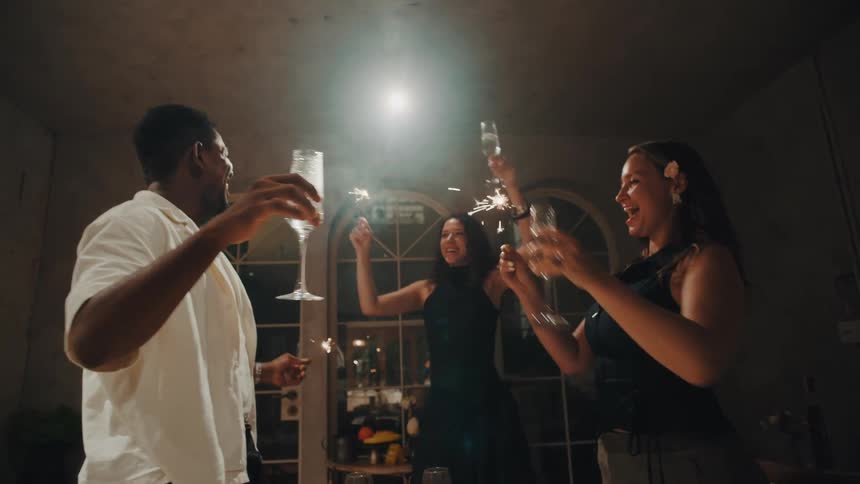 Low angle slow motion shot of three happy, diverse friends toasting with champagne and dancing with sparklers at a festive nighttime party