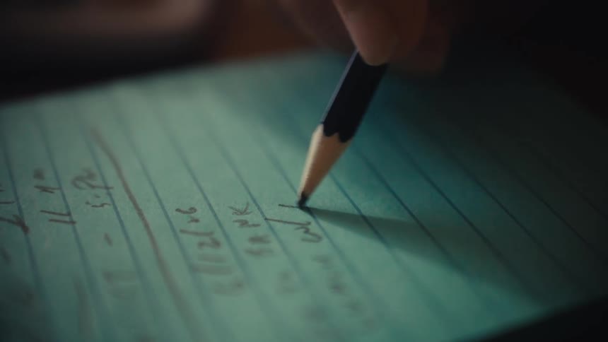 An extreme close-up of a hand using a pencil to write a list on a blue-green lined notepad