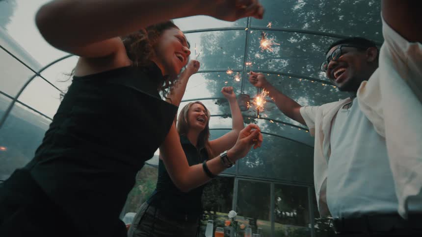 A low-angle slow-motion shot of happy diverse friends dancing and laughing with sparklers at an evening party in a greenhouse