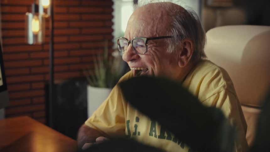 A cheerful elderly US Army veteran laughs heartily while enjoying a video call on his computer at home