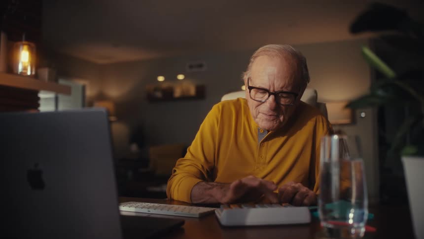 An older man wearing glasses carefully enters numbers on a calculator while sitting at his desk at home in a warmly lit room
