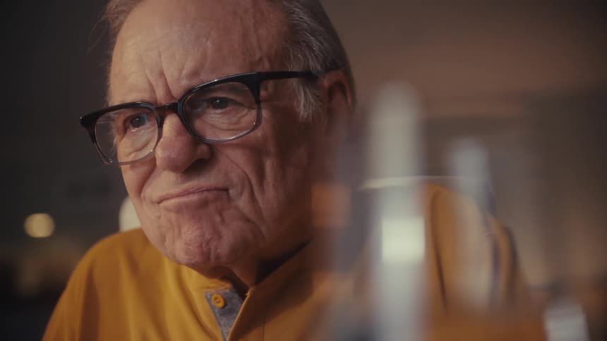 A serious elderly man with glasses looks contemplative in a close-up shot with warm lighting