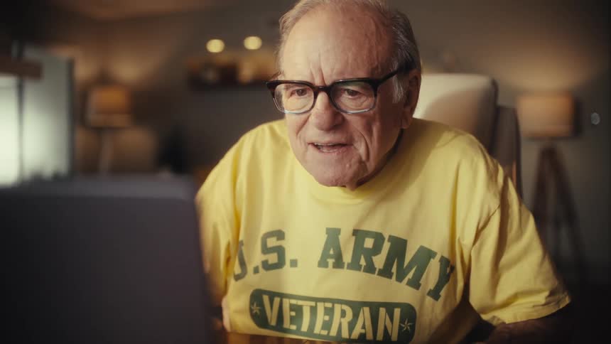 An elderly US Army veteran smiles happily while using his laptop for a video call at his desk