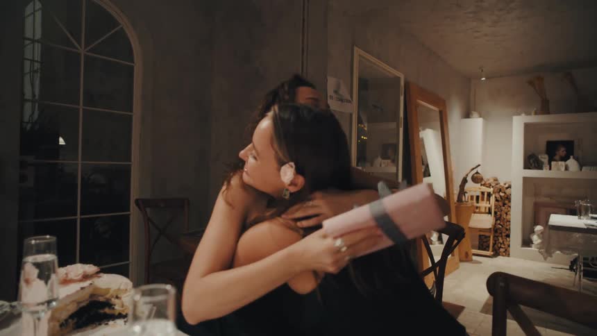 A young woman reacts with surprise and joy upon receiving a birthday gift from her friend at a celebratory dinner, then they embrace warmly