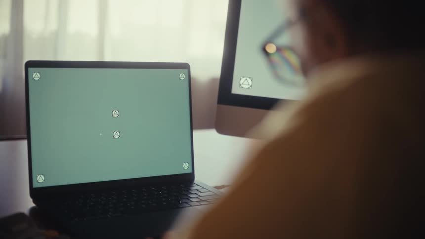 Over-the-shoulder view of a person working on a laptop with a green screen and a second monitor at their desk