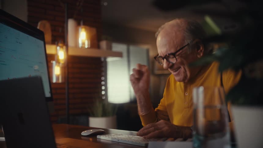 A happy elderly man celebrates a successful outcome while working on his computer in a cozy home office at night