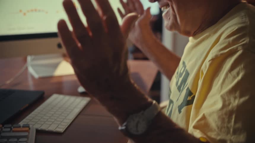 A happy senior man smiles and raises his hands in celebration after seeing positive results on his computer