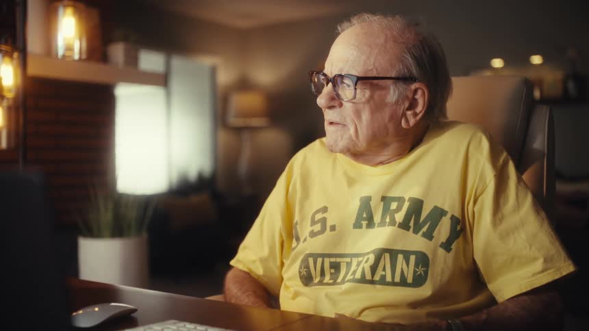An elderly US Army veteran smiles and waves while on a video call on his computer in his home office