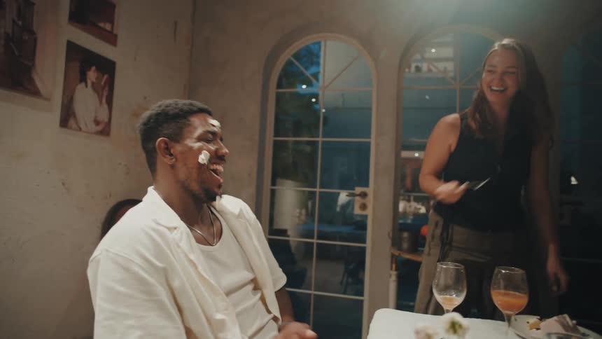 Joyful friends laugh as a woman playfully smears birthday cake on a man's face during a dinner party