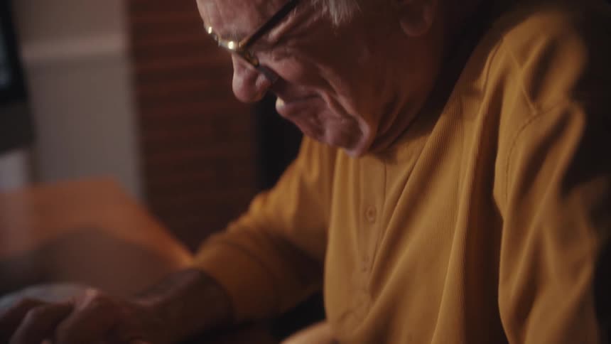 Close up of a happy elderly man in glasses using his computer and typing on the keyboard in a cozy room