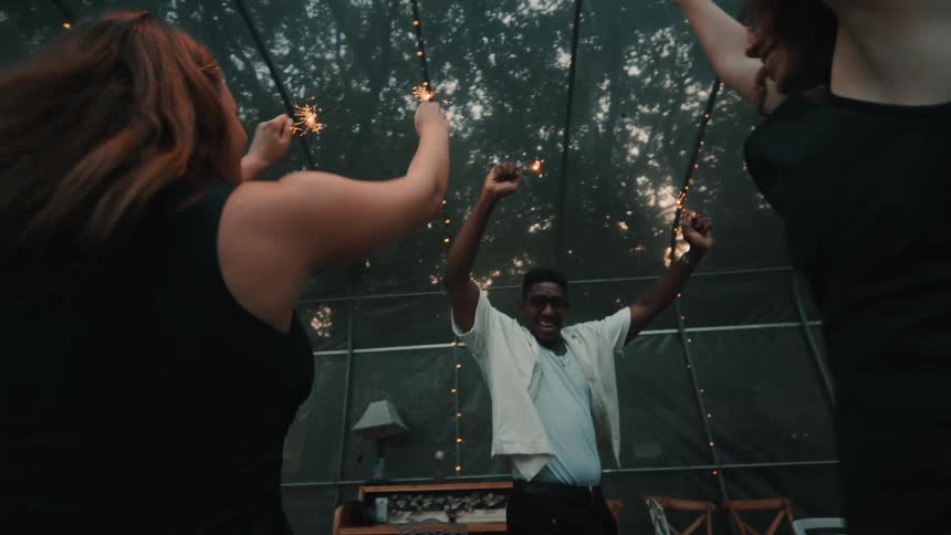 A group of happy, diverse friends dance with sparklers in slow motion at an evening party in a greenhouse