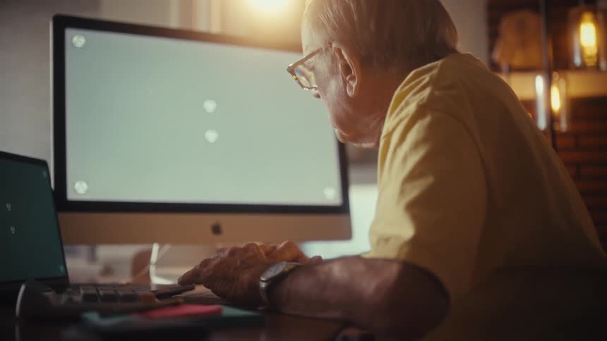 An elderly man in a yellow shirt uses a desktop computer with a green screen in a sunlit home office