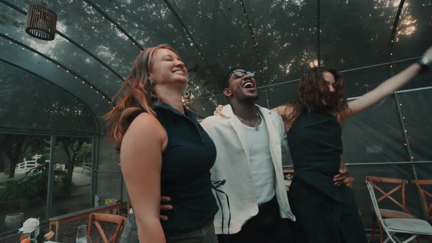 A diverse group of three friends laughs and dances together in slow motion at a festive party inside a beautifully lit greenhouse