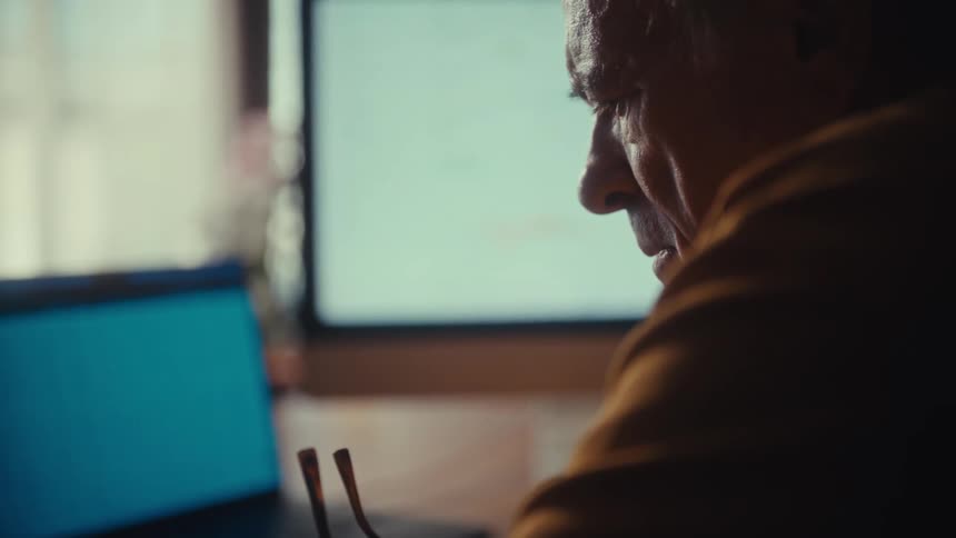 A senior businessman takes off his glasses and contemplates financial charts and spreadsheets on his computer screens in a dimly lit home office
