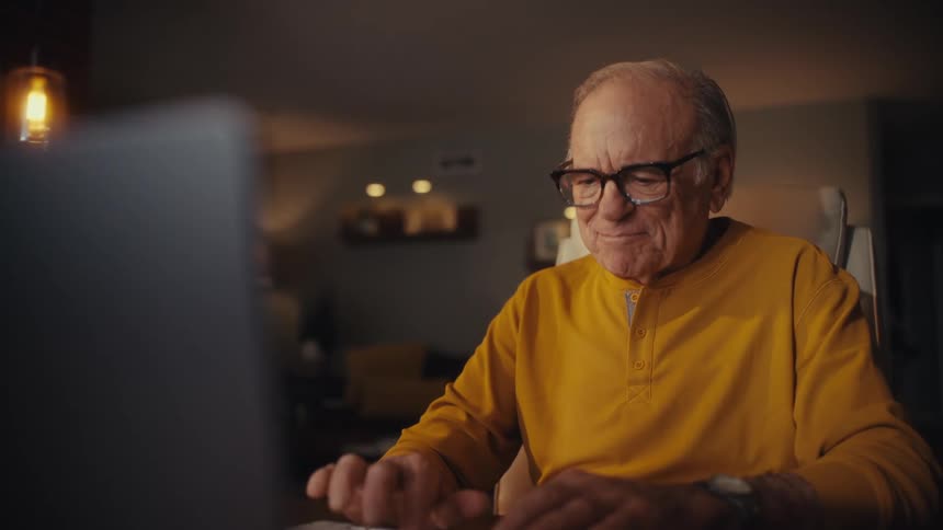 A cheerful elderly man with glasses types on his computer at home in the evening, smiling warmly