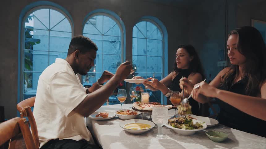 A diverse group of friends shares a meal together, passing plates and enjoying conversation at a dinner party